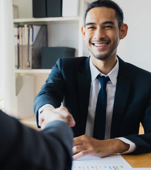 Confident businessman in suit shaking hands at office desk, symbolizing successful partnership.