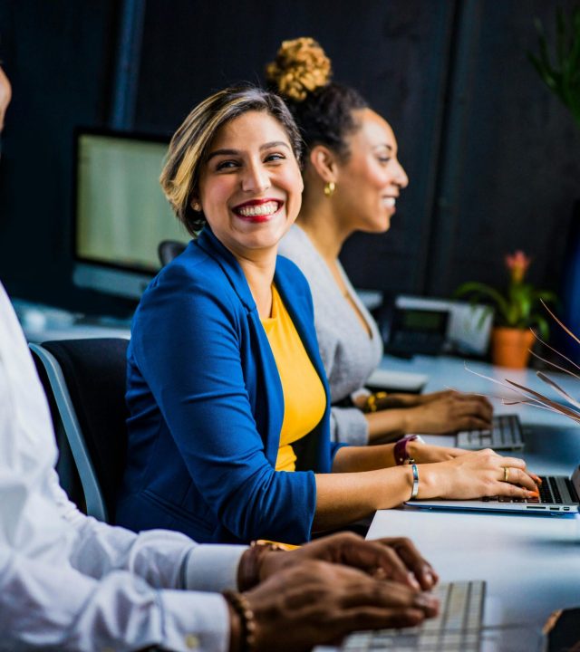 Three diverse professionals working and smiling at office desks, fostering teamwork and collaboration.