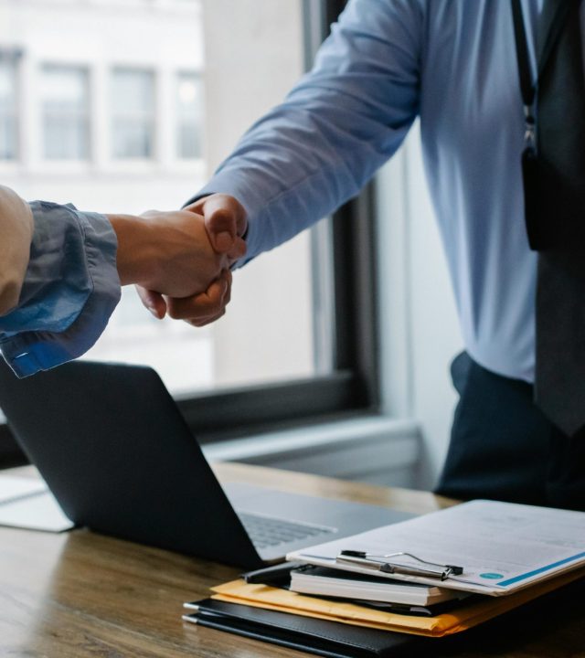 Crop unrecognizable coworkers in formal wear standing at table with laptop and documents while greeting each other before meeting