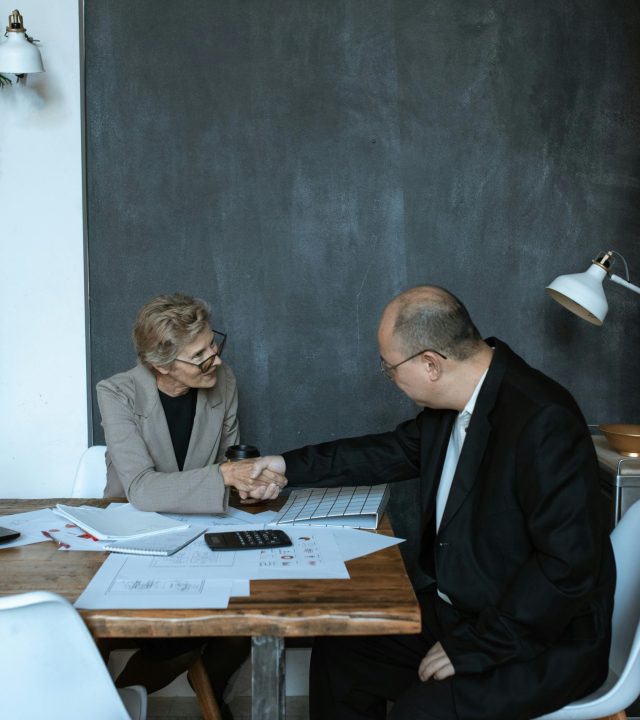 Two professionals engaged in a handshake across a desk in a modern office setting.