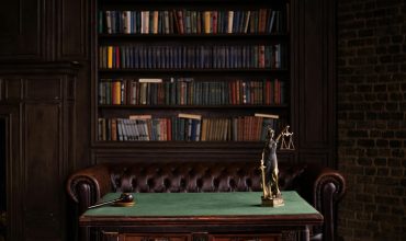 A classic study room with shelves of books, a gavel, and Lady Justice figurine on a green table.