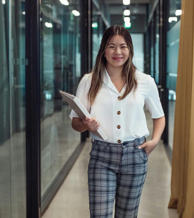 Smiling woman holding documents confidently walking in modern office corridor.