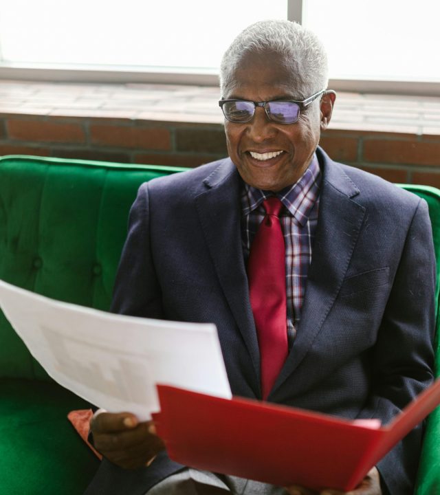 Elderly man in a blue suit sits on a green sofa, reviewing paperwork with a smile.
